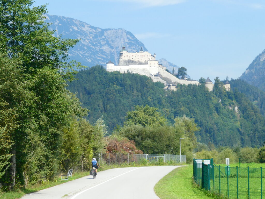 Das Bild zeigt die Burg Hohenwerfen im Hintergrund und im Vordergrund eine Radfahrerin auf der Straße