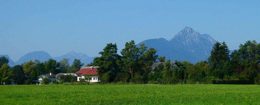 Das Bild zeigt eine Wiese im Vordergrund, ein Haus in der Mitte und einen Berg im Hintergrund