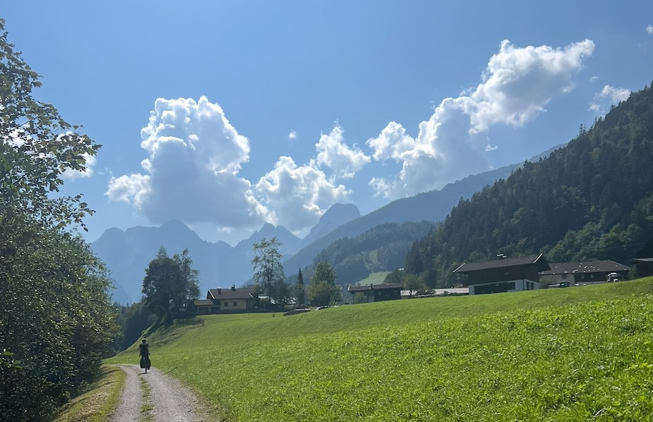 das Bild zeigt einen Radfahrer auf einem geschotterten Radweg, eine Wiese daneben und Berge im Hintergrund